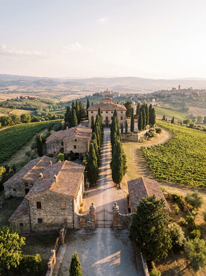 Aerial view of Villa Lucente: stone buildings, iron entrance gates, and a cypress-lined drive among vineyards